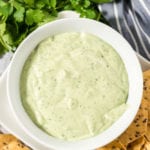 Cilantro avocado dressing in a white bowl surrounded by tortilla chips, cilantro and limes