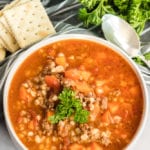 Hamburger soup in a white bowl. A spoon is on the right and a pile of crackers on the left.