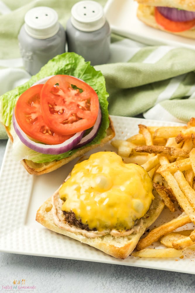 Open face cheeseburger with the burger patty and cheese on one side and lettuce, tomato and onion on the other. A pile of french fries are beside the burger.