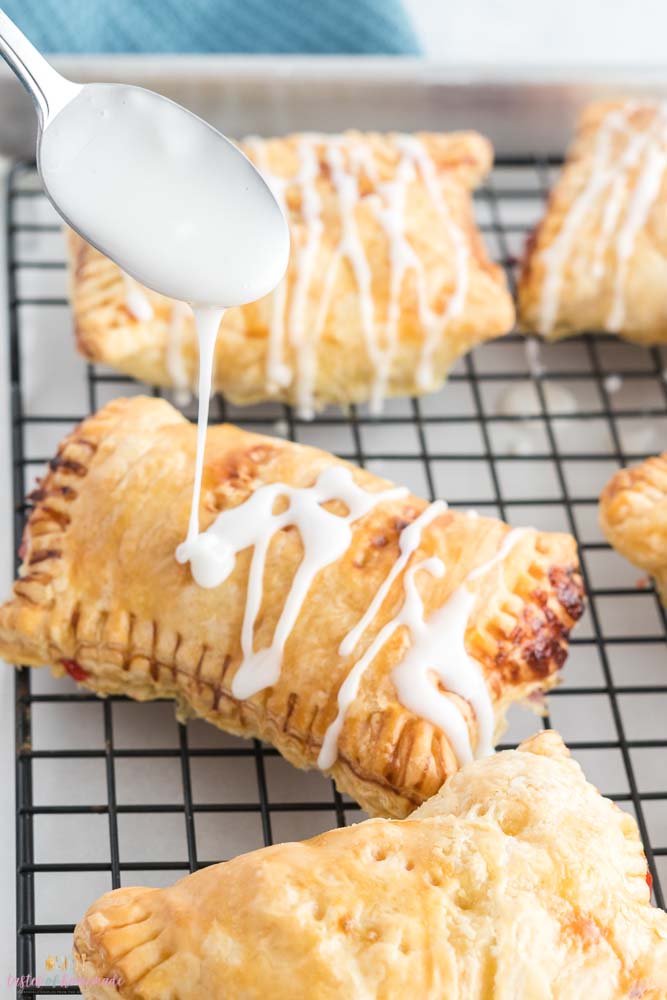 Cherry turnovers on a cooling rack, a spoonful of icing being drizzled on top.