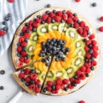 Dessert pizza with raspberries, blueberries, kiwi and oranges. One slice is being removed.