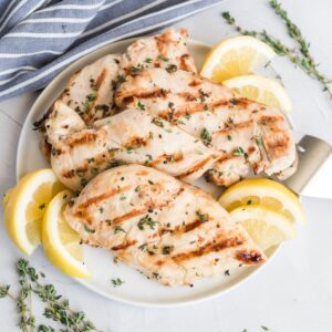 Grilled boneless skinless chicken breast on a plate with lemon wedges and fresh thyme.