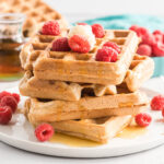 Four waffles stacked on a plate with butter, syrup and fresh raspberries.