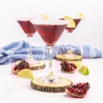 Three pomegranate martinis in stemmed glasses with sugared rim and lime wedges.