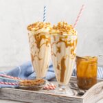 Two Butterscotch milkshakes on a tray topped with whipped cream, butterscotch sauce and crunchy toffee bits.