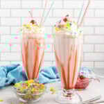 Two milkshakes in tall glasses with strawberry syrup, whipped cream and topped with fruity pebbles.