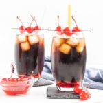 Two cocktails made with cola and grenadine in tall glasses garnished with a cocktail stick and three cherries.