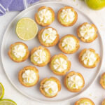 Mini lime curd tarts topped with whipped cream on a platter with lime slices.