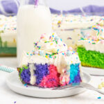 A slice of cake coloured with purple, pink and blue swirls, topped with white frosting and rainbow sprinkles.