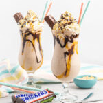 Two milkshakes in tall glasses with caramel and chocolate sauce topped with whipped cream, chopped peanuts and half a snickers bar.