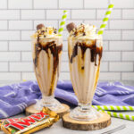 Two chocolate milkshakes in tall glasses with chocolate and caramel sauce, topped with whipped cream, half a twix bar and a straw.