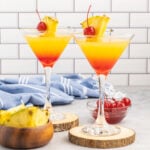 Two martinis layers with grenadine and pineapple juice in martini glasses and garnished with cherries and pineapple wedge.
