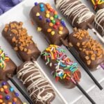 Chocolate covered marshmallows decorated with rainbow sprinkles on cake pop sticks on a white plate.
