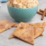 A pile of cinnamon pita chips on a table with a bowl of pumpkin dip beside them.