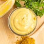Small jar of mayonnaise with a slice of lemon and spoonful of curry seasoning on a cutting board.