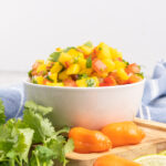 Fresh mango salsa in a white bowl with whole habanero peppers and a bunch of cilantro.