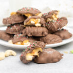 Two cashew turtles on a table with a bite out of one showing gooey caramel centre and a plate of turtles in the background.