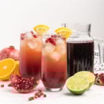 Two sparkling pomegranate drinks in tall glasses garnished with orange and lime wedge.