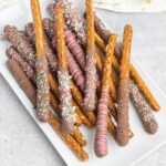 A platter of chocolate dipped pretzel rods decorated with candy sprinkles and chocolate drizzle.