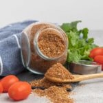 A small jar of taco seasoning with a spoonful of seasoning on a table with cherry tomatoes and cilantro leaves.