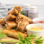Cheesburger egg rolls on a wooden board with dishes of mustard and ketchup.