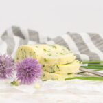 Pats of chive butter sitting on a piece of parchment paper surrounded by fresh chives and chive blossoms.