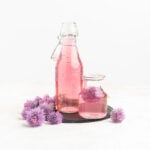 A bottle of pink coloured vinegar on a slate plate surrounded by chive blossoms.