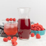 A bottle of raspberry syrup, a glass of fizzy drink made with syrup, and a dish of fresh raspberries.