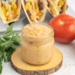 A jar of burger sauce, with burger tacos and a tomato in the background.