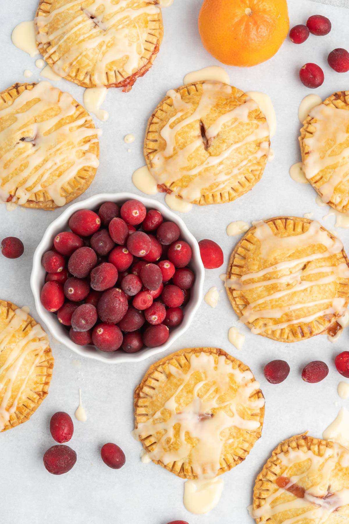 Round handpies with icing drizzle on a table with a bowl of cranberries and two oranges.