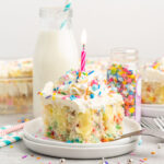 A slice of birthday cake with colorful sprinkles and a lit candle.