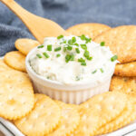 Chive cream cheese with a wooden spreader in a small bowl with crackers on a plate.