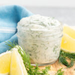 Dill mayonnaise in a small jar with lemon wedges around it.