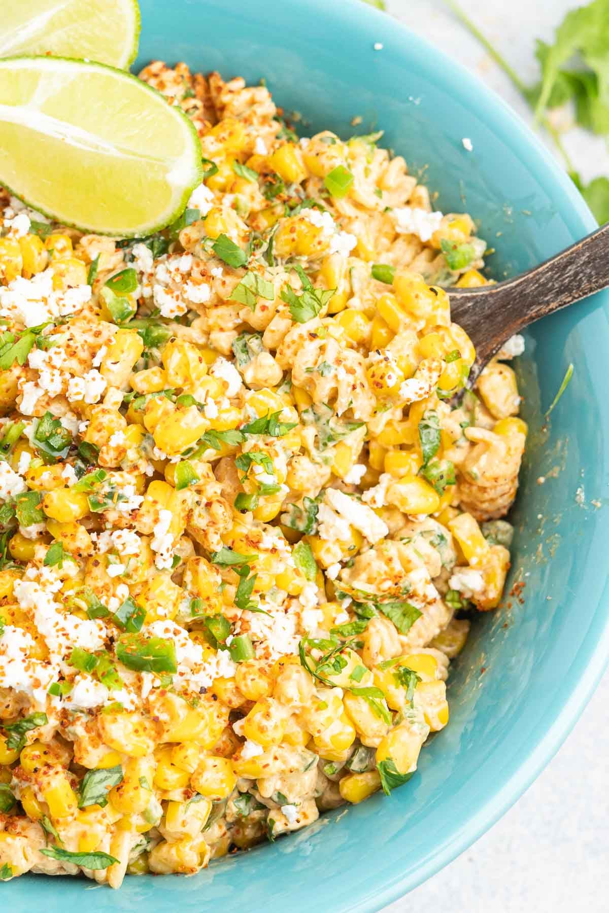 Pasta salad with corn and crumbled cotija cheese in a bowl garnished with lime wedges. 