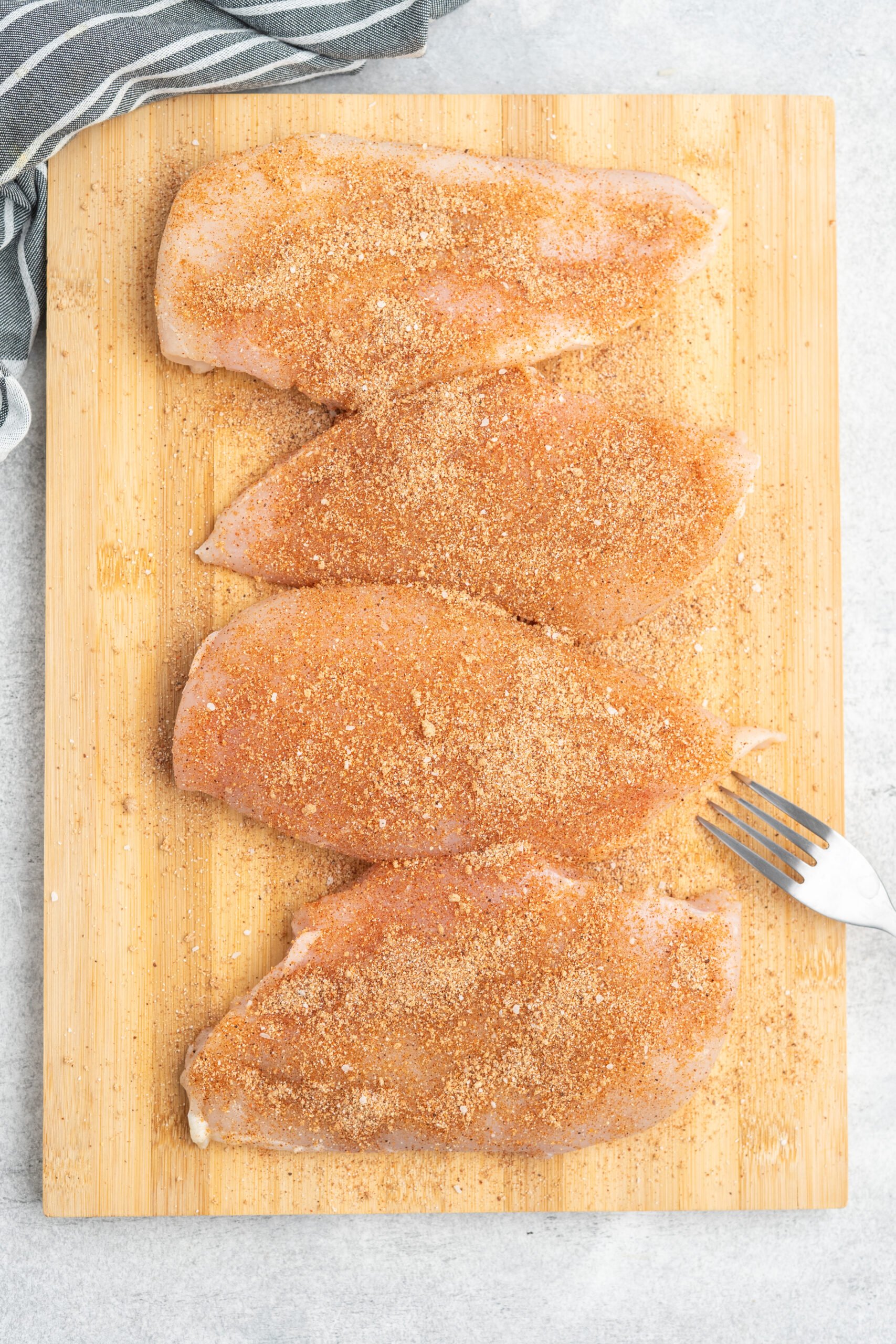 Four raw boneless, skinless chicken breasts seasoned with BBQ seasoning on a cutting board. 