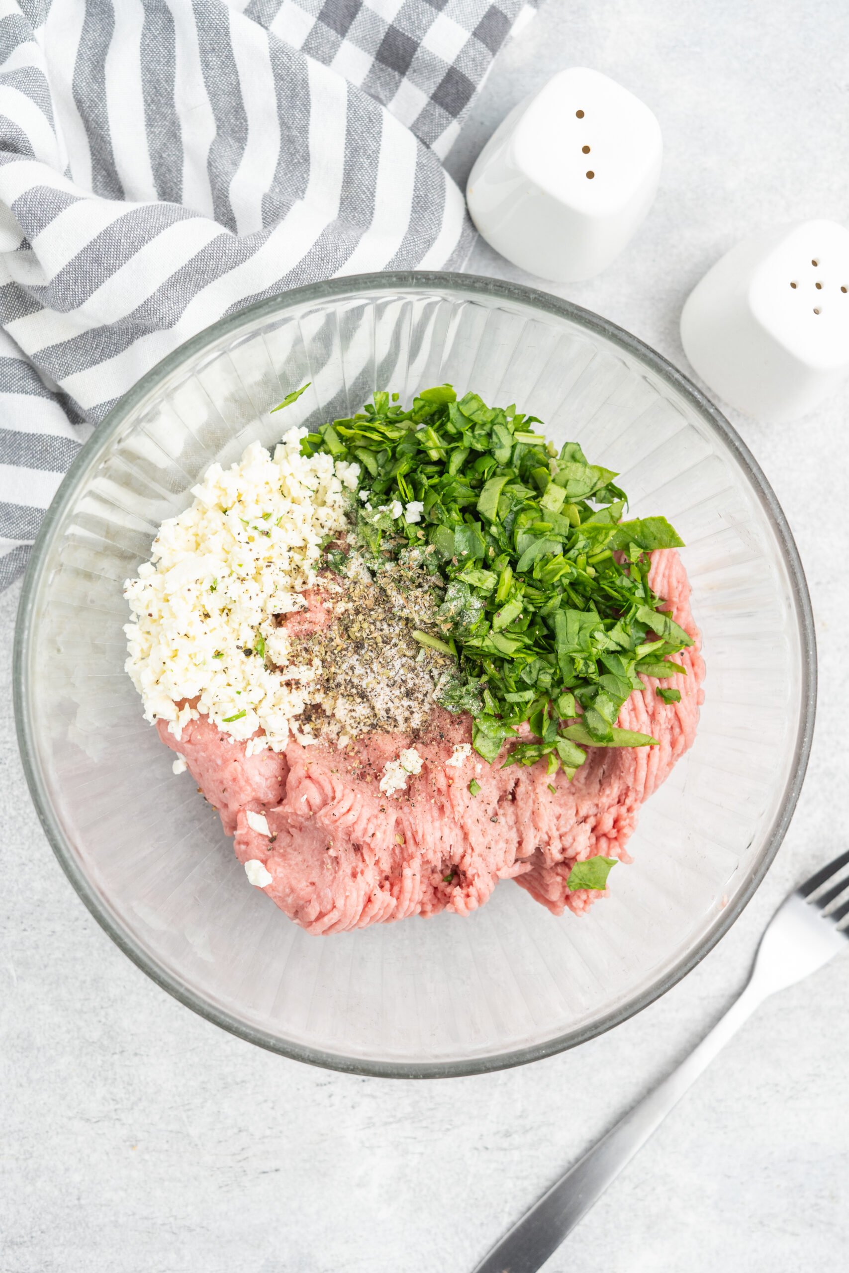 A glass mixing bowl with ground turkey, chopped spinach, crumbled feta, and spices.